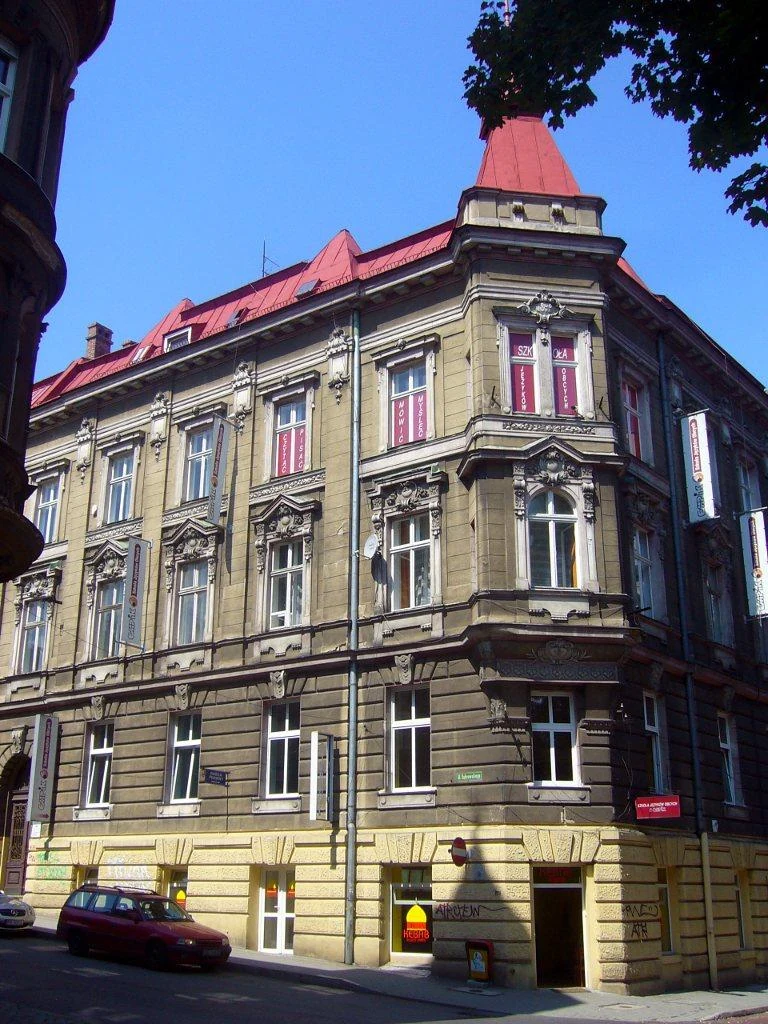Narożna kamienica przy Dąbrowskiego 6 i Mickiewicza 25, z wykuszem, ozdobną elewacją i czerwonym dachem.